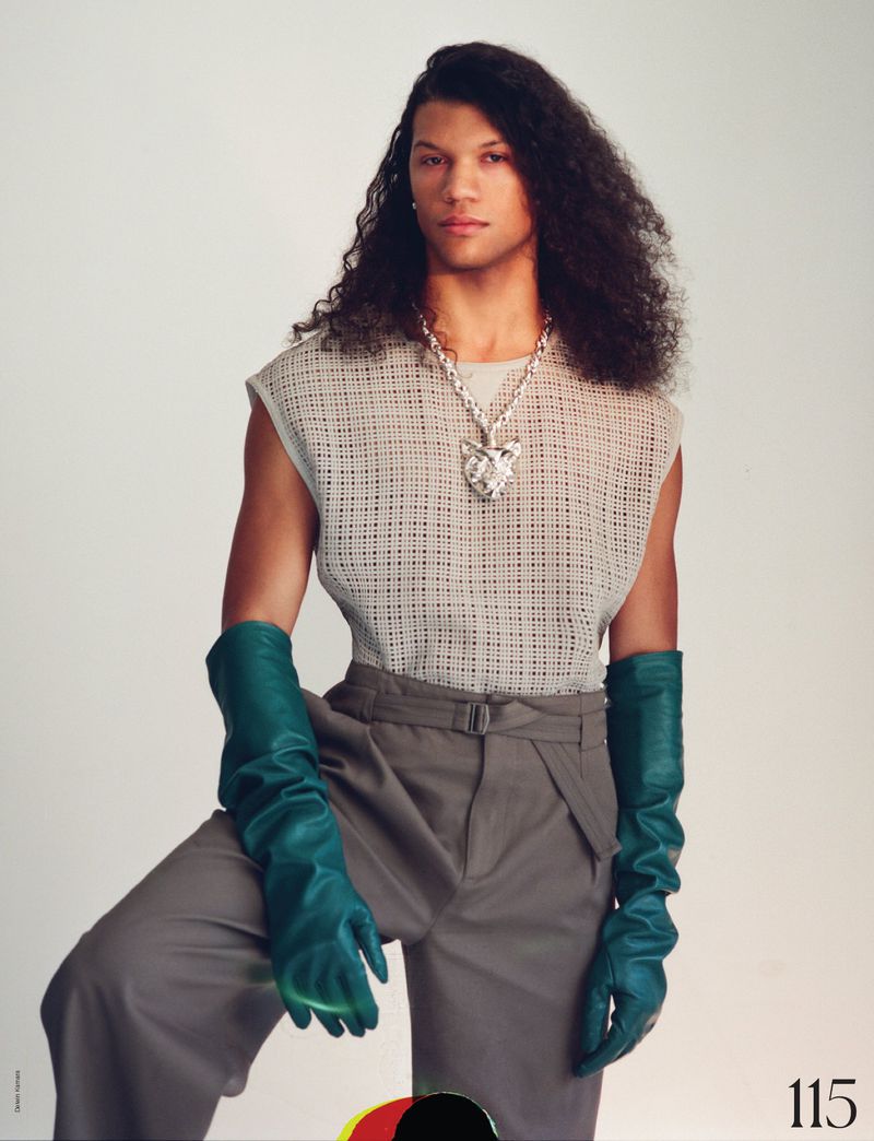 Adult male with long, wavy brown hair poses wearing a gray sleeveless mesh top, gray pants, large pendant necklace, and teal gloves.