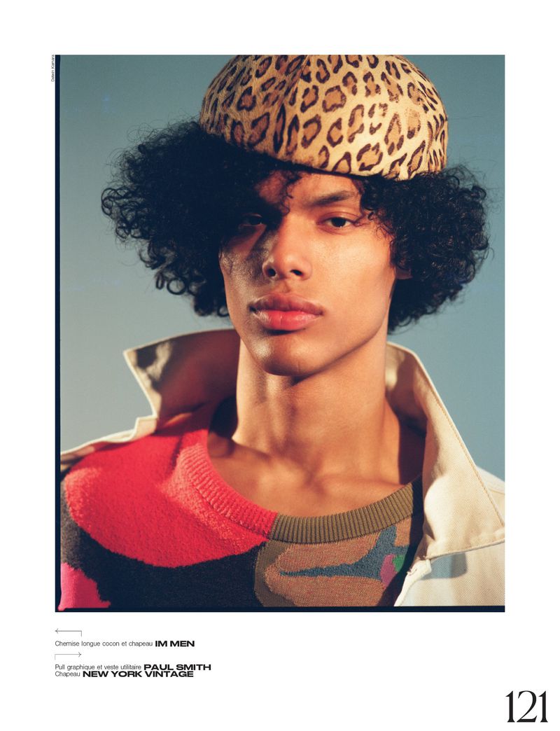 Close-up of a dark-skinned male model with an afro wearing a cheetah-print hat, colorful sweater, and beige trench coat against a blue background.