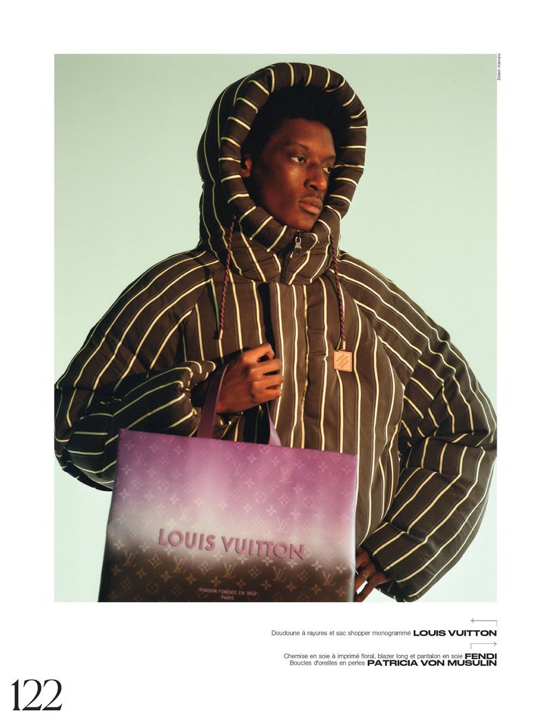 Black male model poses with Louis Vuitton shopping bag, wearing striped hooded puffer jacket in a studio with a light teal background.