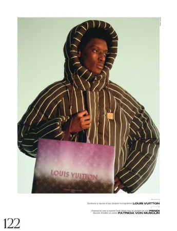 Black male model poses with Louis Vuitton shopping bag, wearing striped hooded puffer jacket in a studio with a light teal background.