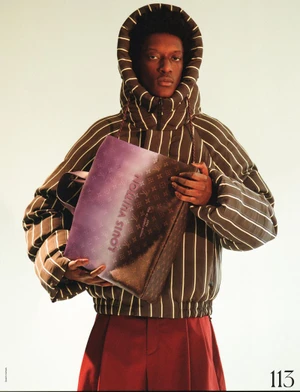 A male model wearing a striped puffer hooded jacket and holding a monogram handbag against a neutral white seamless background.