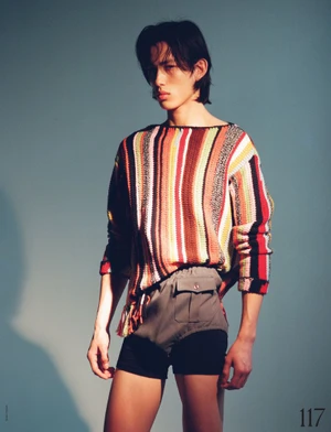 Young male model wearing a striped knit sweater and dark shorts against a blue-grey studio background for French Magazine.