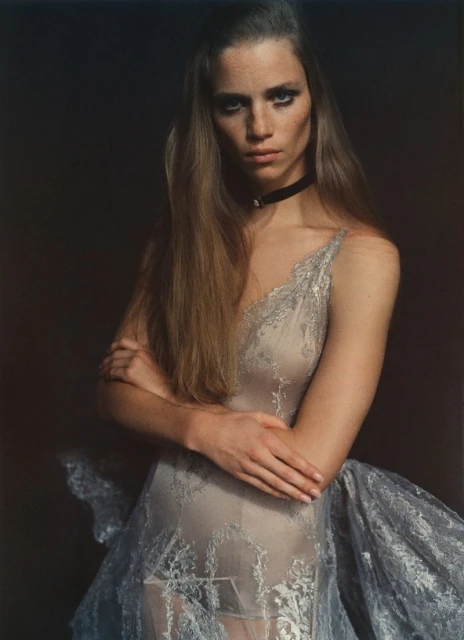 Portrait of an adult woman with long brown hair, wearing a lace dress and choker necklace, standing in a dark photo studio.