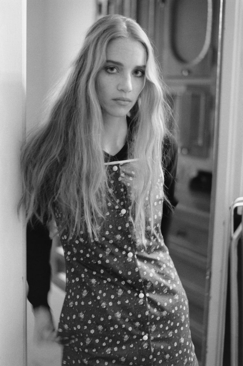 Black and white portrait of a young adult female model with long hair, wearing a polka dot dress, standing indoors against a wall.