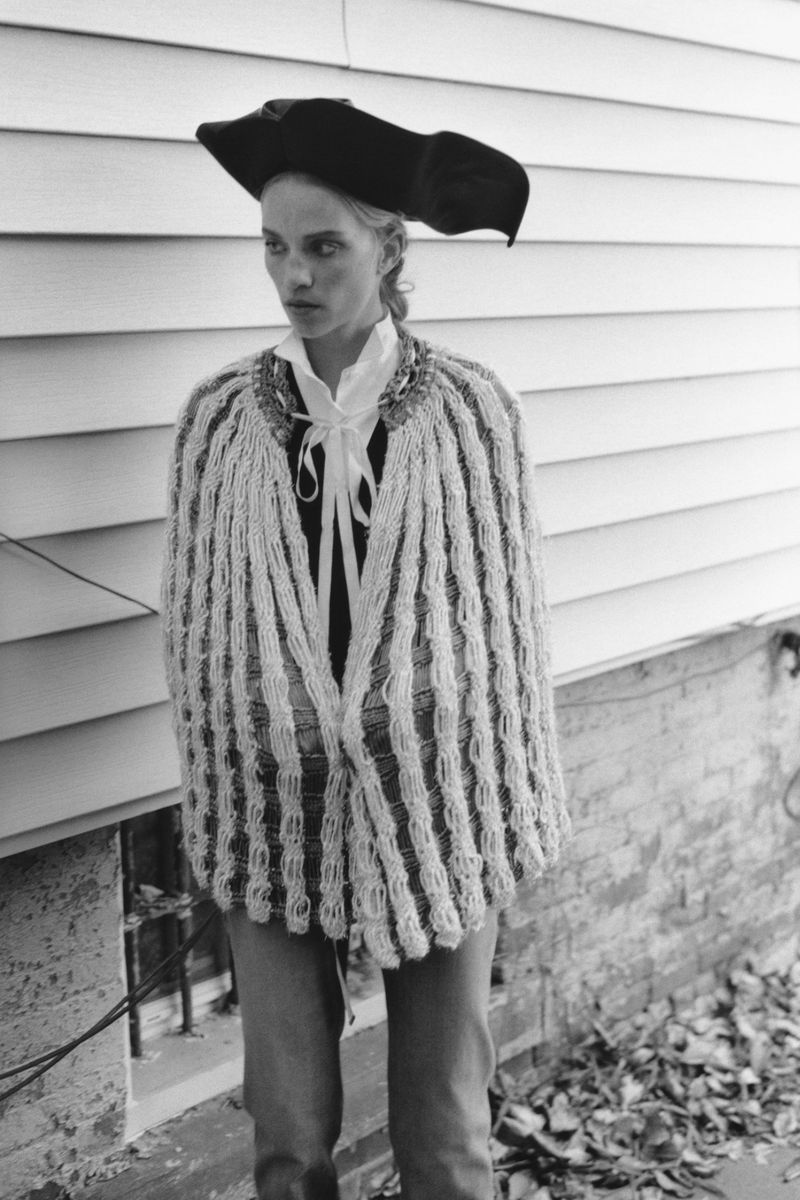 Young woman in historical costume poses against siding outdoors. Knitted shawl, tricorn hat, and white collared shirt are visible.