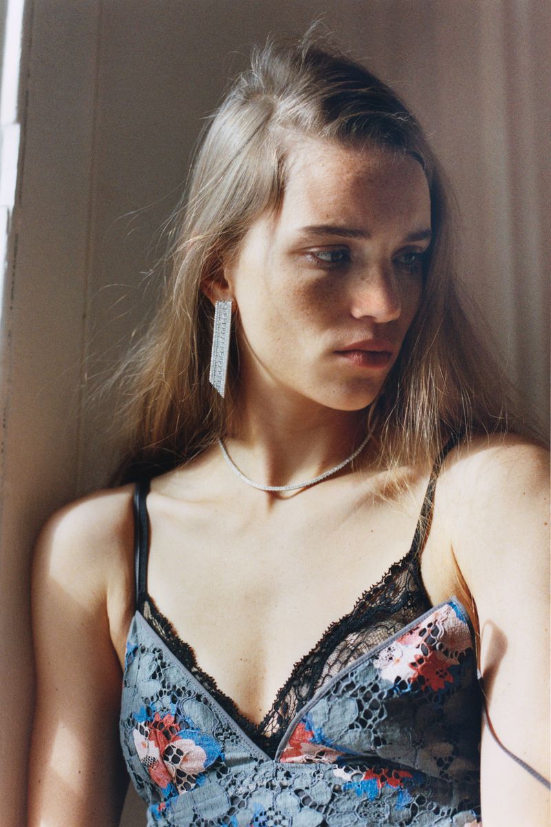 Young woman with long brown hair poses in floral top with black lace trim. She is wearing long silver earrings and a silver necklace.
