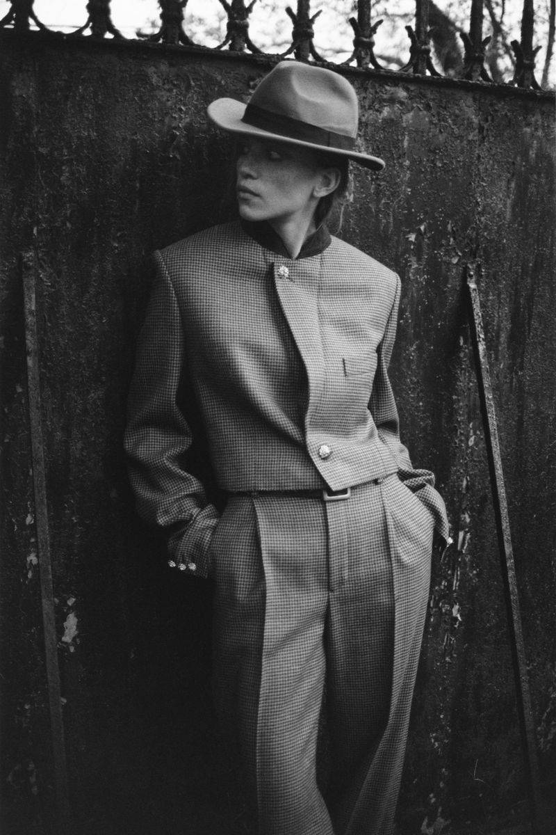 Monochrome portrait of a stylish adult leaning against a rustic wall in a houndstooth suit and fedora hat looking left.