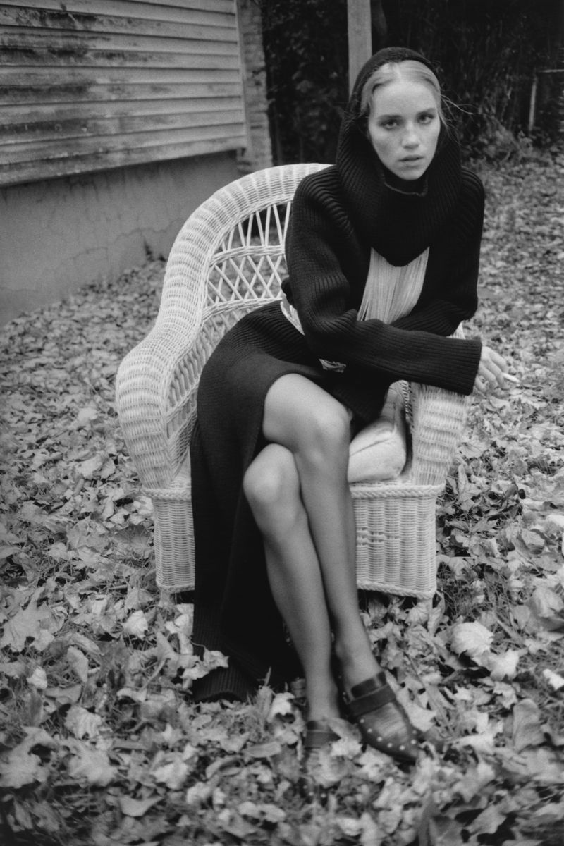 Black and white portrait of a woman seated in a wicker chair outdoors among fallen leaves, wearing a dark sweater dress.