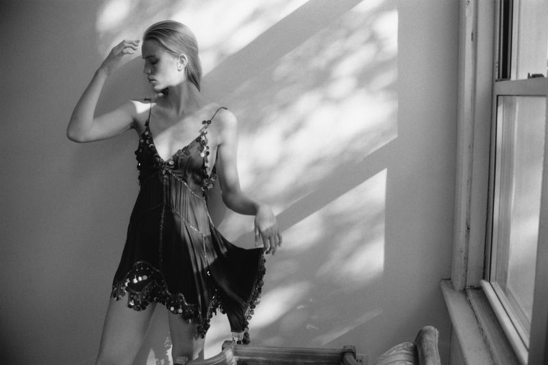 Model Rebecca Leigh Longendyke in a dark slip dress standing beside a window with dappled sunlight on the wall for Self Service.