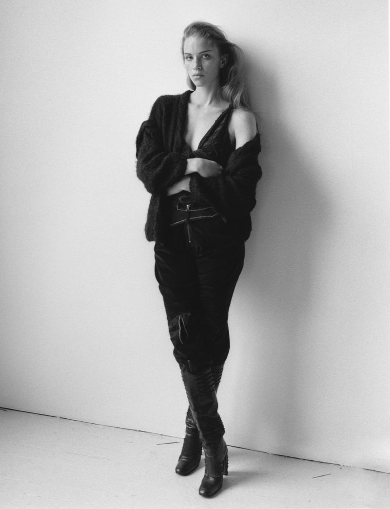 A woman stands against a white wall wearing a black cardigan, matching top, black trousers and tall black leather boots.