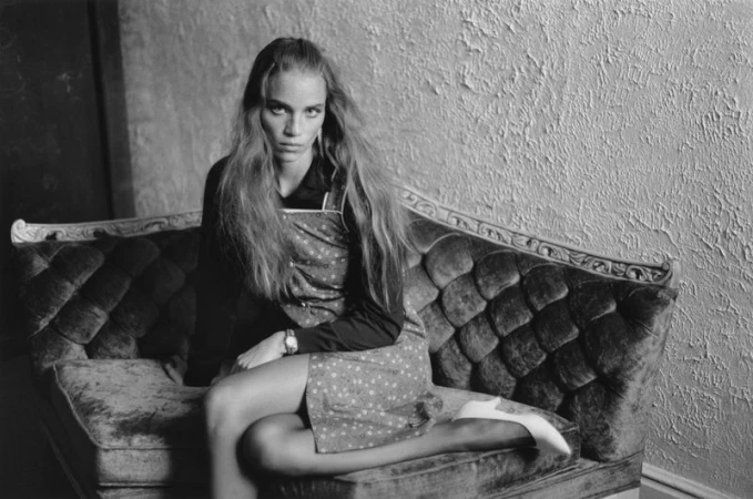 Black and white portrait of an adult woman with long hair sitting on a tufted couch in front of a textured wall.  
