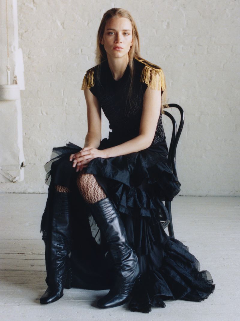 A fashionable woman poses while seated, wearing a black dress with fringe shoulder detail and leather boots against a neutral backdrop.