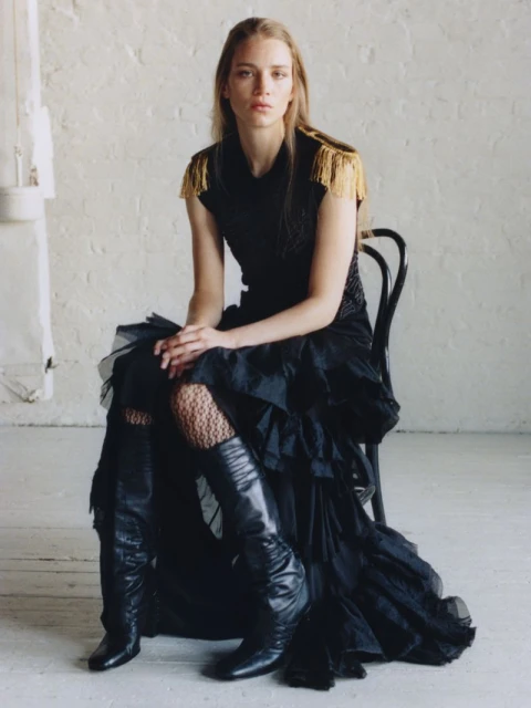 A fashionable woman poses while seated, wearing a black dress with fringe shoulder detail and leather boots against a neutral backdrop.