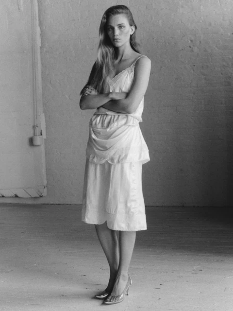 Full-length portrait in black and white of a slim adult woman with long hair, wearing a light-colored dress and heels, arms crossed.