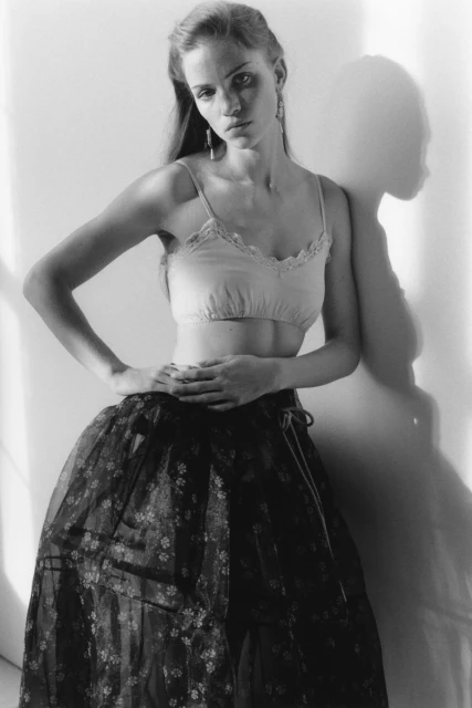 Woman in bralette and full skirt poses against a wall, casting a shadow. Studio shot in black and white with soft lighting.
