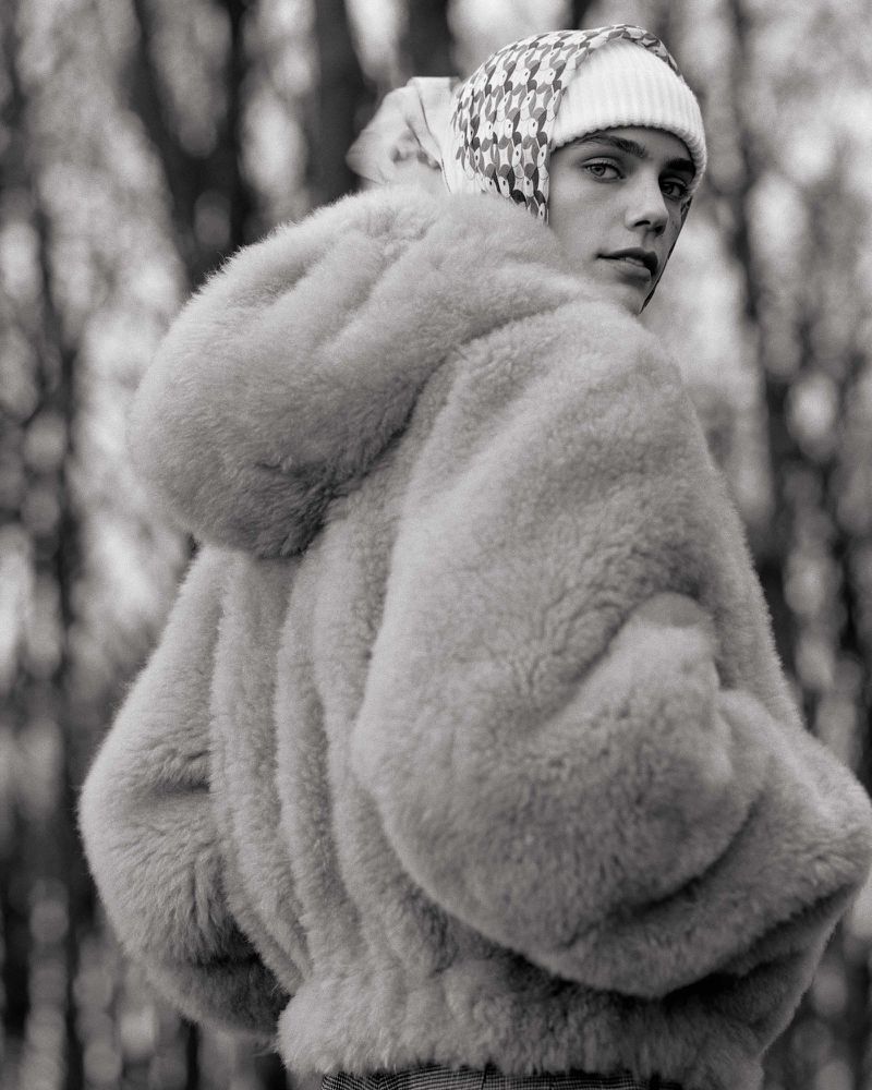 A person in a beige fur coat with a patterned hat stands in a wooded area looking over their shoulder in this black and white portrait.