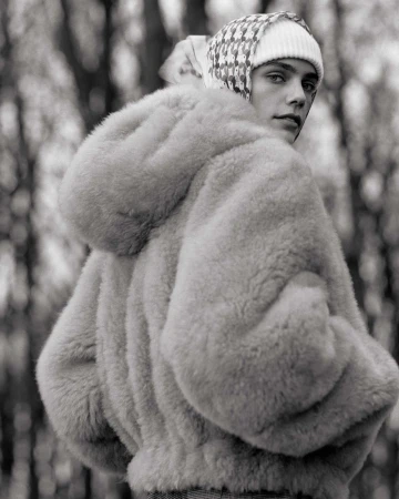 A person in a beige fur coat with a patterned hat stands in a wooded area looking over their shoulder in this black and white portrait.