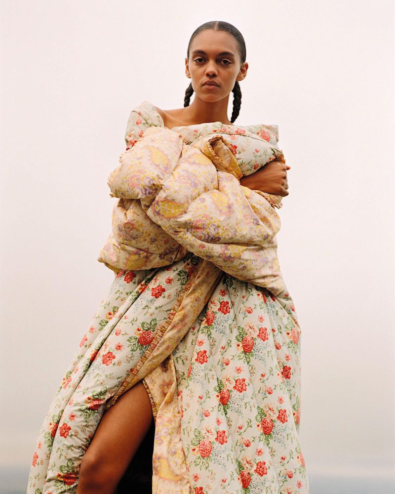 Fashionable woman in floral puffer gown, poses outdoors with a neutral expression. Puffed sleeves, thigh-high slit, clear sky backdrop.