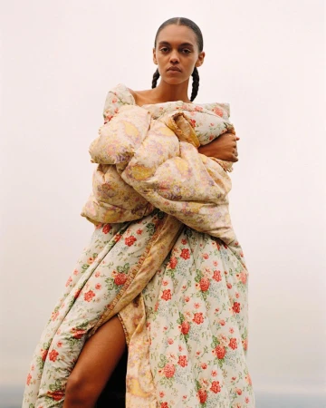Fashionable woman in floral puffer gown, poses outdoors with a neutral expression. Puffed sleeves, thigh-high slit, clear sky backdrop.