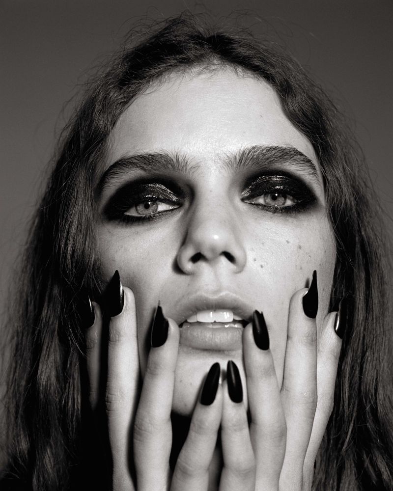 Dramatic black and white beauty shot of a woman with dark eye makeup and long black nails posing with her hands near her mouth.