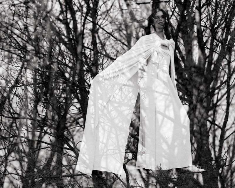 Monochrome image of a woman in a white gown standing among tree branches, creating a stark and ethereal fashion portrait in nature.