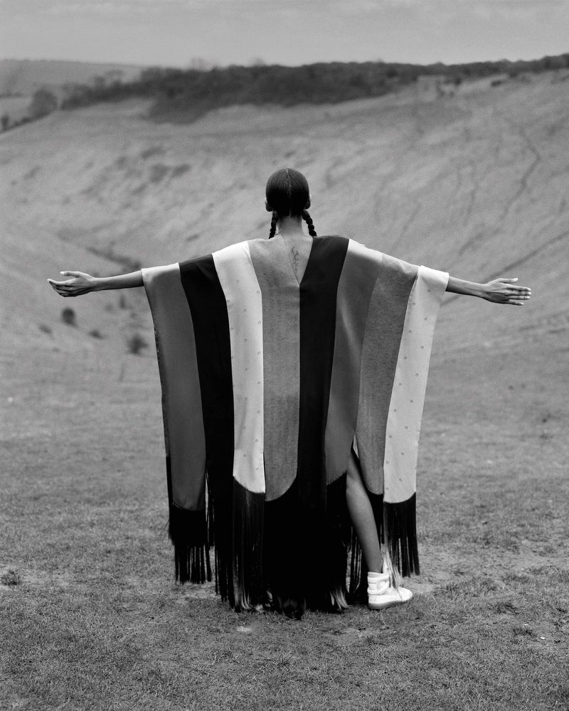 Back view of a woman standing in a field, wearing a colorful poncho with fringe, with rolling hills visible in the background.
