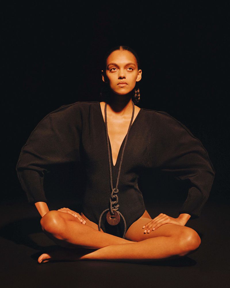 Attractive female model in black bodysuit and necklace poses in lotus position against dark background in fashion studio shot.