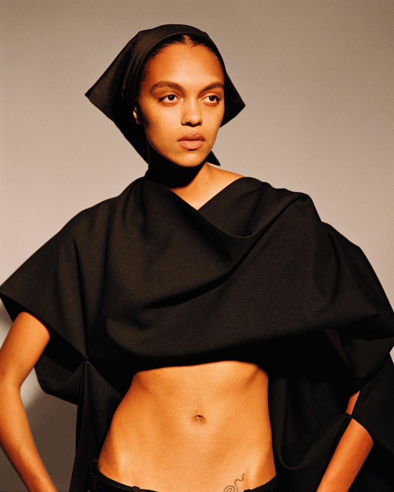 Woman in a black headscarf and draped garment poses in a studio setting. Minimalist fashion shot with soft, diffused lighting and a neutral backdrop.
