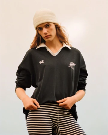 Fashionable young woman in a black rugby shirt, white hat, and striped pants strikes a pose in a brightly lit outdoor scene.