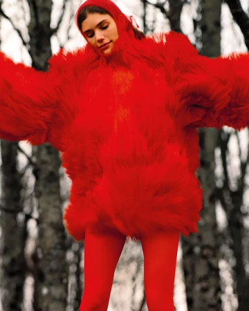 Woman in striking red costume in snowy forest. The image features bold color contrast against the trees, full-body shot.
