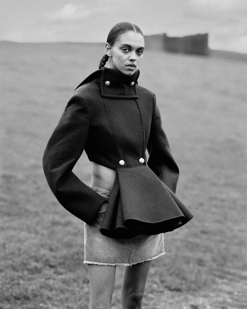 Woman in a cropped black jacket and denim skirt posing outdoors in a field. Fashion shot in black and white with a neutral expression.
