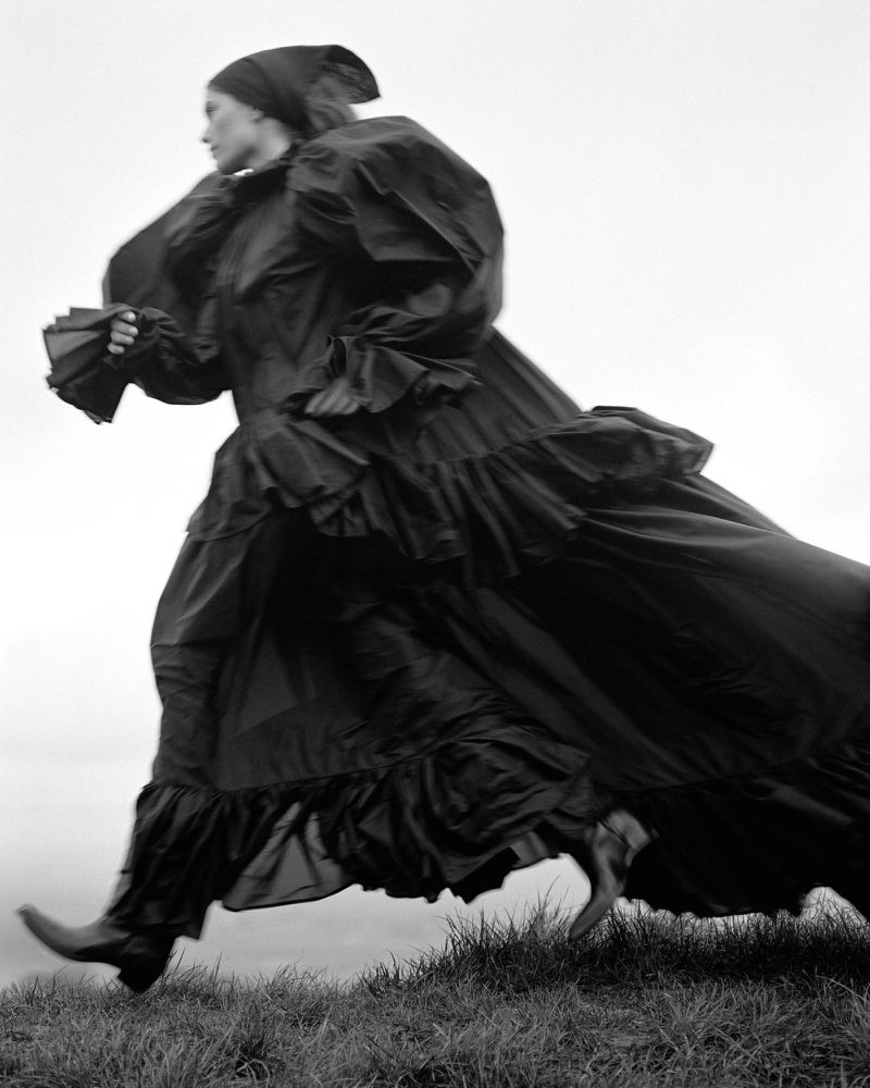 Fashionable woman in motion wearing a black gown and headscarf, running across a grassy hill against a bright, monochromatic background.