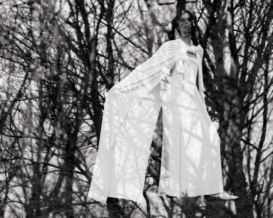 Model in flowing white cape and trousers posed among bare winter tree branches in a black and white editorial photograph.