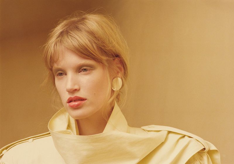 Close-up studio portrait of a blonde woman with soft makeup, wearing a light yellow jacket and gold earrings against a cream backdrop.