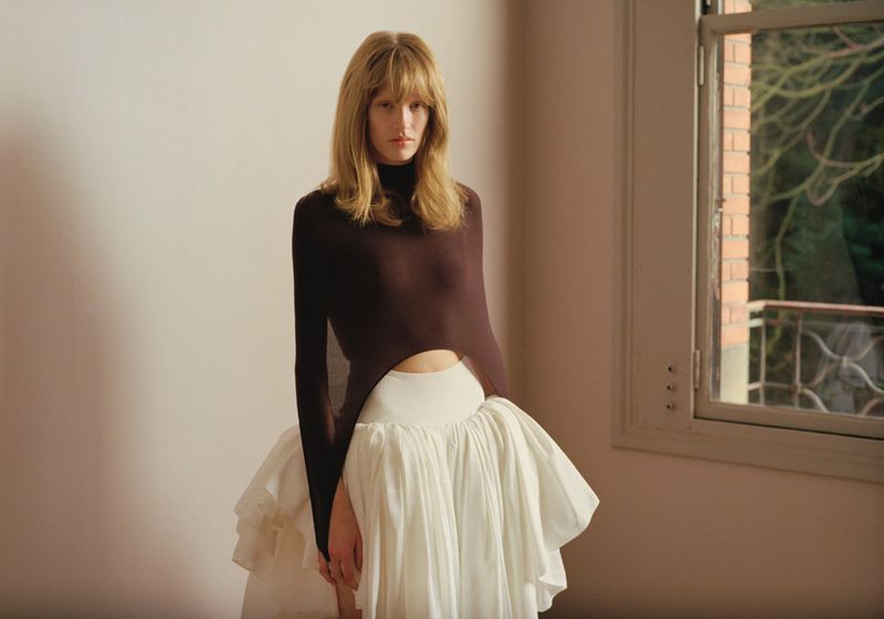 A slender woman with bangs poses indoors in a long-sleeved brown top and a voluminous white skirt, looking directly at the camera.
