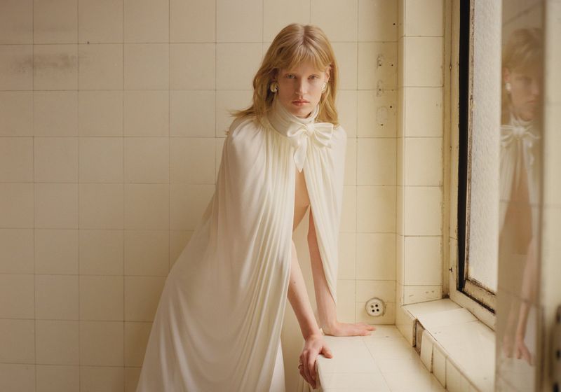 Elegant blonde woman in a white gown with a large bow, posed in a tiled indoor setting with natural light, fashion-forward and refined.