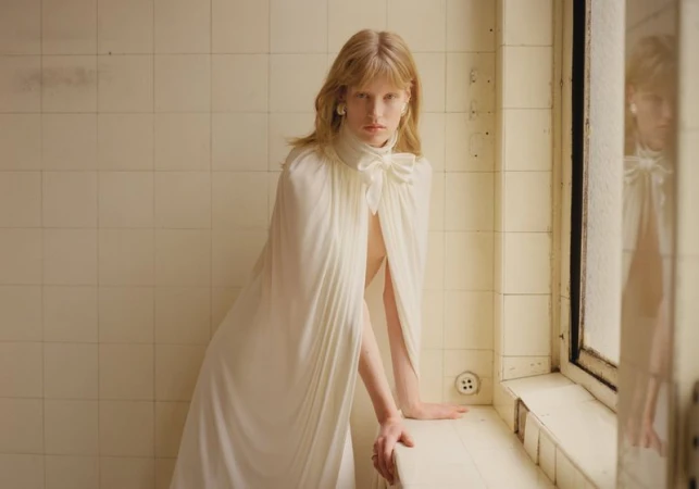 Elegant blonde woman in a white gown with a large bow, posed in a tiled indoor setting with natural light, fashion-forward and refined.