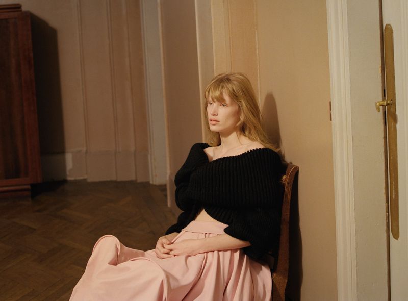 Adult woman with blond hair sits on a chair in a pink outfit and black sweater in an indoor setting with neutral colors and natural light.