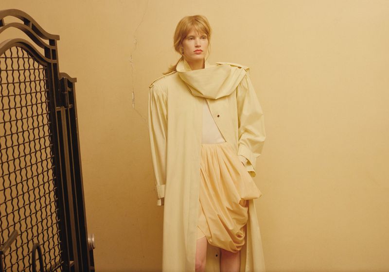 Woman in a yellow trench coat standing against a yellow wall with a metal gate, posing for a fashion editorial shot, studio lighting.