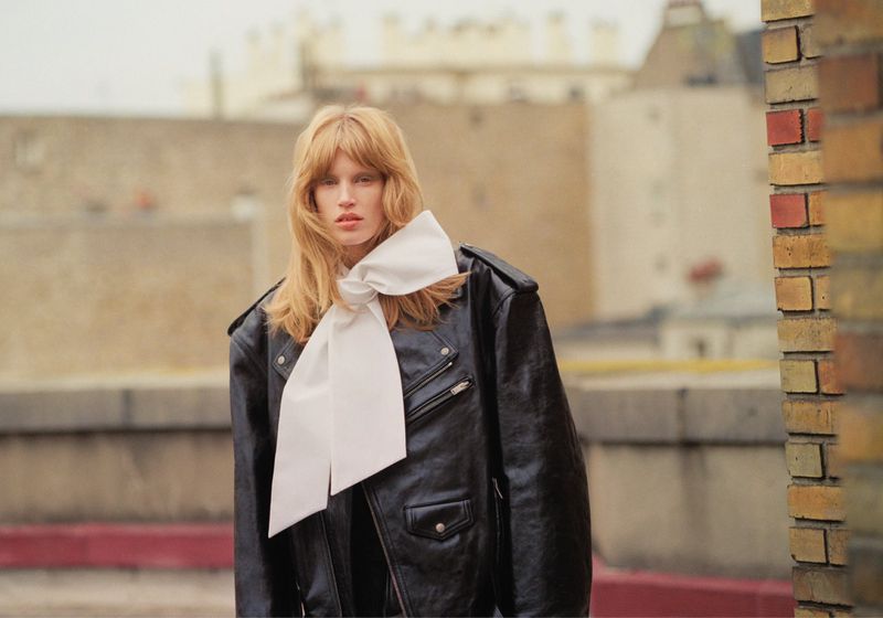 A fair-skinned woman with flowing red hair in a black leather jacket and white satin bow posing on a rooftop with neutral city buildings.