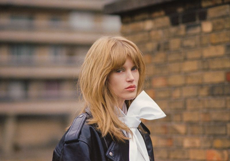 Model with blonde hair wearing a black leather jacket and a large white bow against a brick wall background