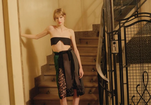 A woman poses on a staircase, wearing a black tube top and a draped skirt with lace details in a warm, indoor setting.