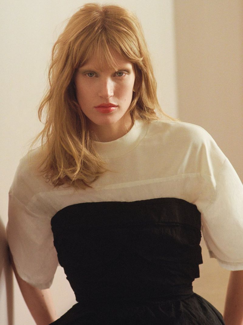 Portrait of a female model wearing a white top and a black strapless bodice against a neutral wall interior.