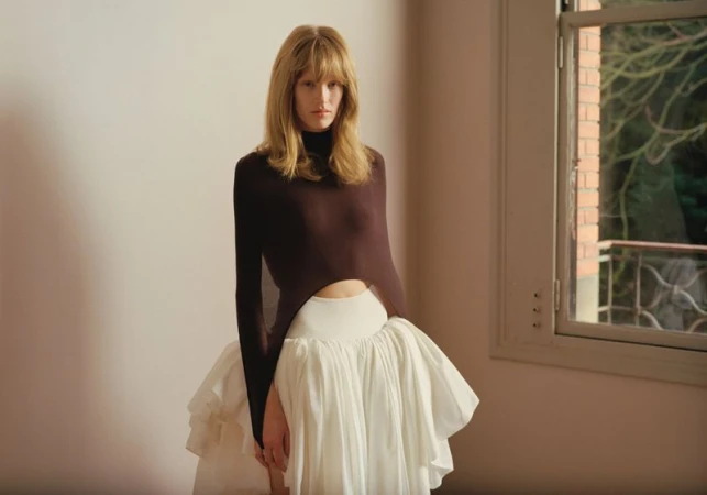 Model with blonde hair wearing a dark fitted long-sleeve top and voluminous white skirt standing in a room