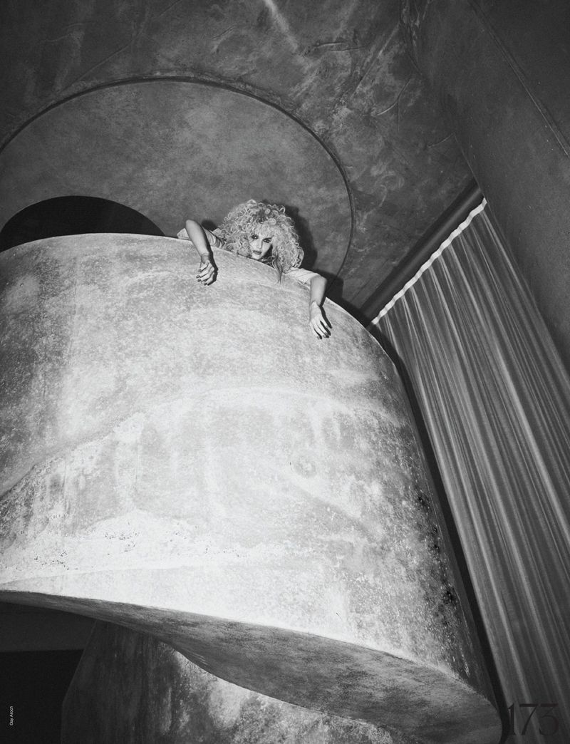 Monochrome portrait of an adult female with curly blonde hair hanging from a concrete structure in an indoor environment with curtains.