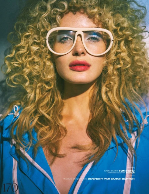 Close-up portrait of a blonde adult female wearing glasses and a blue jacket. The image features a studio setting with soft lighting.