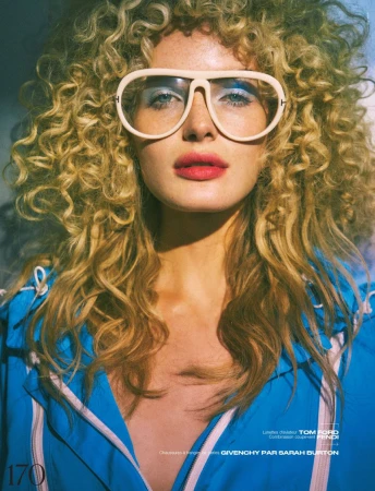 Close-up portrait of a blonde adult female wearing glasses and a blue jacket. The image features a studio setting with soft lighting.