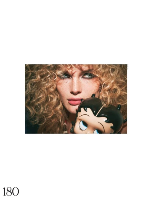 Fashion portrait of a woman with curly blonde hair holding a Betty Boop bag, against a white background in a studio setting.