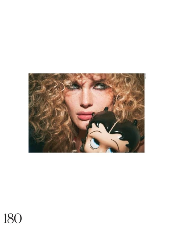 Fashion portrait of a woman with curly blonde hair holding a Betty Boop bag, against a white background in a studio setting.