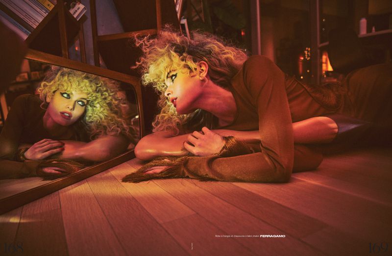 Woman with blonde curly hair posing with a mirror on the floor in indoor setting. Reflections and warm lighting create mood.